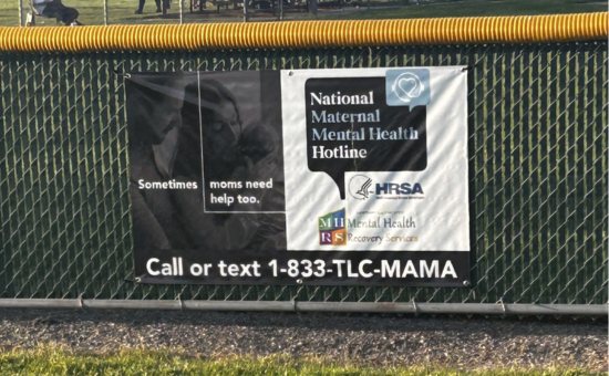 Black and white banner promoting the National Maternal Mental Health Hotlines hanging on a baseball fence