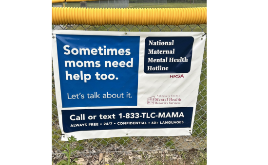 Blue and white banner promoting the National Maternal Mental Health Hotlines hanging on a baseball fence